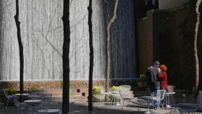 Man and woman watch waterfall in Paley Pocket Park in Manhattan, New York City. Park is filled with white tables and chairs.