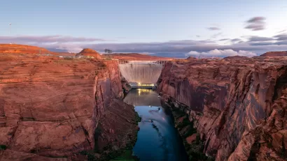 View of Glen Canyon Dam with vertical red stone cliffs on either side, Colorado River at bottom, and dusk sky in background.