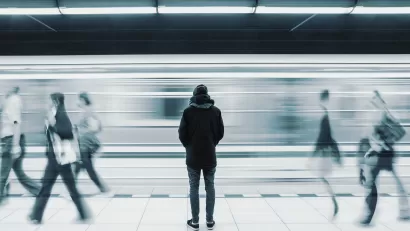 Monotone photo of man waiting on subway platform with blurry train passing by and other passengers walking by
