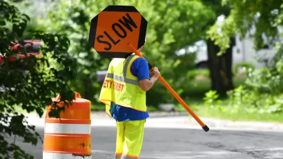 Worker in yellow safety suit holding up orange SLOW sign on road