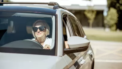 Senior woman wearing sunglasses sitting in the driver's seat of a silver car.