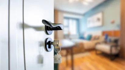Close-up on door handle with door key inserted and blurred furnished room visible beyond.