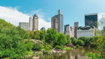 View of Central Park lake with people sitting on lakeside rocks and NYC high-rises in background.