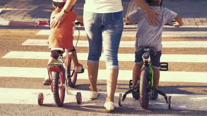 Close-up from back of woman walking on crosswalk with two small children on either side of her riding bicycles with training wheels.