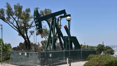 Green oil well pump jack surrounded by fence on a hill overlooking neighborhood.