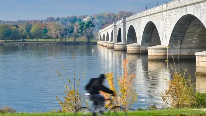Person on bike on path next to Memorial Bridge in Washington, D.C. with fall foliage in background across river.