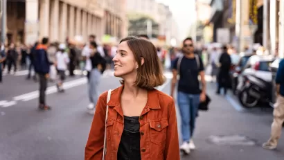 Brown-haired woman in an orange jacket walking down crowded street smiling
