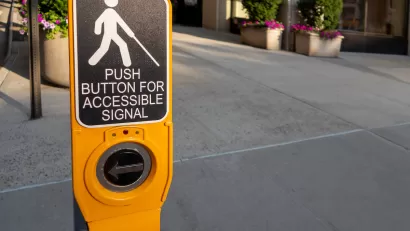 Close-up of yellow pedestrian crossing signal button with text "Push button for accessible signal" and diagram of person holding cane, indicating blindness.