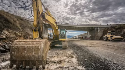 Excavator parked on side of highway under construction with overpass bridge in background.