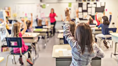 View from back of classroom of elementary school children at desks with raised hands engaged in class.