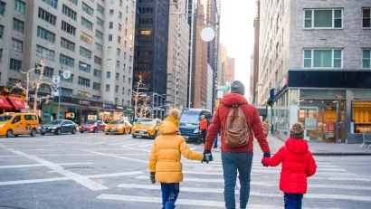 Adult holding hands of two children, all wearing winter coats, in crosswalk in New York City during holidays with trees decorated with lights in background.