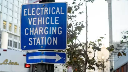 Close-up on blue and white "Electric vehicle charging station" sign with arrow pointing right on pole on city street.
