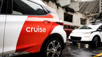 White and red Cruise autonomous car on San Francisco street with concrete Brutalist-style building in background on grey, rainy day.