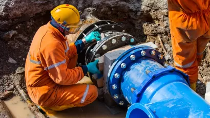 Worker in orange suit and hard hat working on large blue water pipe in ground.