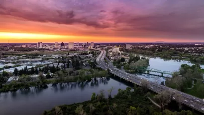 Aerial view of freeway crossing river in Sacramento, California with colorful sunset sky in background.