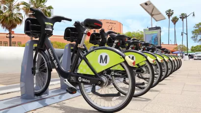 Row of black and green Los Angeles Metro bike share bikes at station with red building with clock and'Los Angeles' lettering in background.