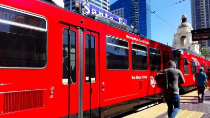 Red San Diego Trolley light rail train at station with passengers walking on platform and Spanish-style dome building in background.