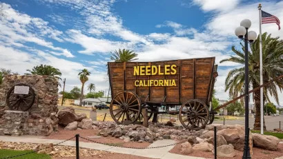 Sign for Needles, California installed on wooden wagon with residential neighborhood visible in background.