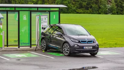 Small grey sedan charging at outdoor electric car charging station with green kiosk and green forest in background in Norway.