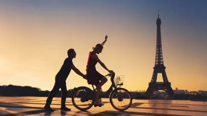 Silhouette of man holding on to back of bicycle ridden by woman with Eiffel Tower in background.