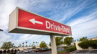 Close-up of red sign with white "Drive-Thru" text and arrow pointing left against partly cloudy blue sky.