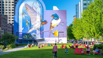 People enjoying a sunny day on a green lawn with pink lawn chairs and colorful bird mural in background in Boston, Massachusetts.