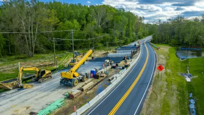 Aerial view of road construction with yellow heavy equipment parked next to two-lane asphalt road against green forest and blue sky