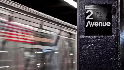 Close-up of 2 Avenue sign on column in NYC subway station with train passing by in background.