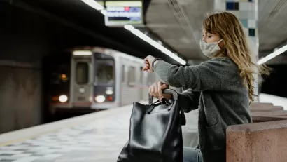 Woman with long hair wearing Covid mask sitting on underground train station bench looking at her watch as subway train approaches in background at Hollywood/Western station in Los Angeles, California.