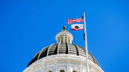 Close-up of top of California state capitol dome with U.S. and California flags flying and blue sky in background.