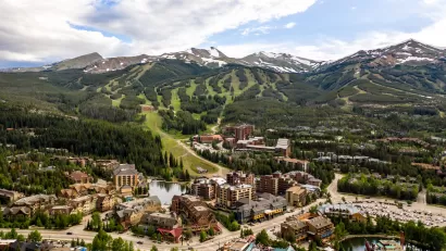 An aerial view of the town of Breckenridge, Colorado, in the foreground with lots of businesses, houses, and a ski resort, with green treed mountains in the backround and taller snow-capped mountains in the distance.