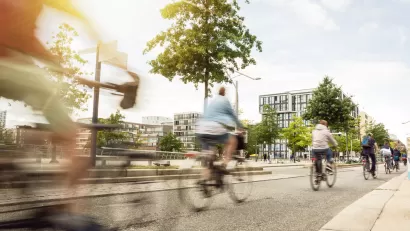Line of five cyclists bike away from camera in wide protected bike lane on the right side of a street, with low-rise residential buildings in the background.