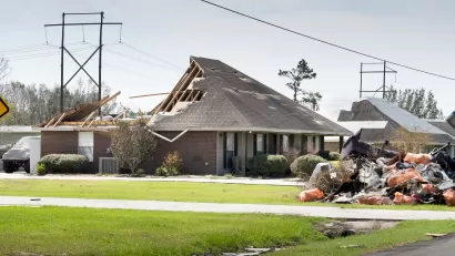 Exterior side view of a single-story brick home missing back half of roof and a debris pile in the front yard.