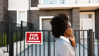 Woman realtor or home buyer client wearing blazer walking past urban condo with red and white For Sale sign while talking on phone.