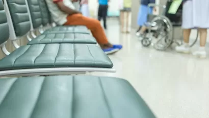 Close-up of blurred empty vinyl chairs with person sitting in background and wheelchair visible in hospital waiting room.