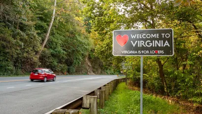 Small red car driving on forested road passing "Welcome to Virginia" sign.