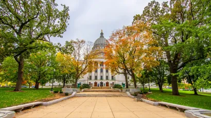 Green trees, some with fall colors, line a walkway in front of the Wisconsin state capitol dome in Madison, WI.