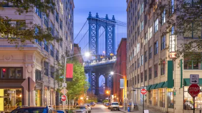 View of Brooklyn Bridge at night from Brooklyn alley with multi-story buildings and parked cars.