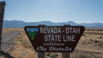 Roadside sign for Nevada Utah State Line "Entering Ely District" with Bureau of Land Management logo with desert mountains in background.