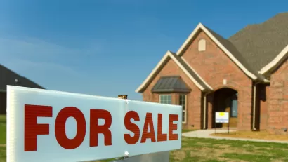 Close-up of white sign with red FOR SALE letters and blurred brick single-family home in background.