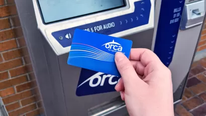 Close-up of person's hand holding blue ORCA transit fare payment card in front of fare payment machine.