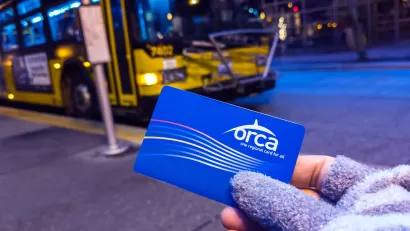 Close-up of gloved hand holding blue Orca transit card with blue and yellow bus in background