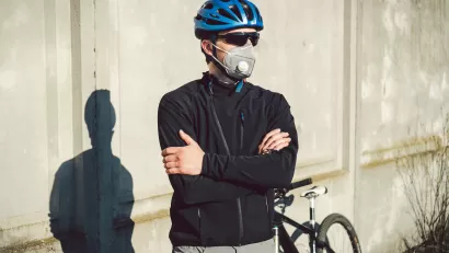 Male cyclists wearing helmet, sunglasses, and N95 mask stands with arms crossed in front of his bike