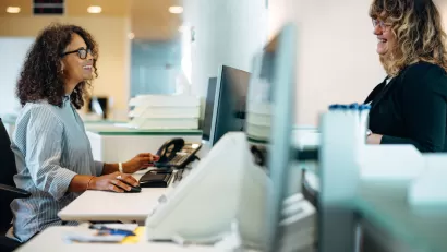 Two women on either side of reception desk talking to each other with row of computers between them.