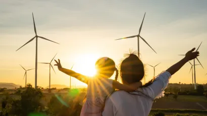 Adult holding young child facing away from camera looking at wind turbines sillhouetted against the sunset.