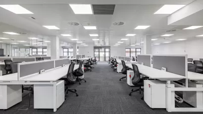 Large empty open-plan office with rows of white desks and black chairs under bright fluorescent lighting