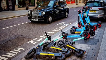Group of e-scooters messily parked on street in London with black cab in background.