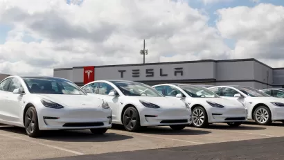 Row of white Tesla electric cars in parking lot with dealership building with Tesla logo in background.