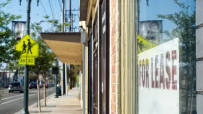 Close-up of For Lease sign in window of shop storefront with city sidewalk and vintage street light in background.