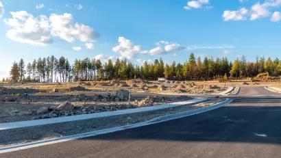 Empty subdivision lots with new roads and sidewalks complete and line of evergreen forest in the immediate background under a bright blue sky with fluffy white clouds..